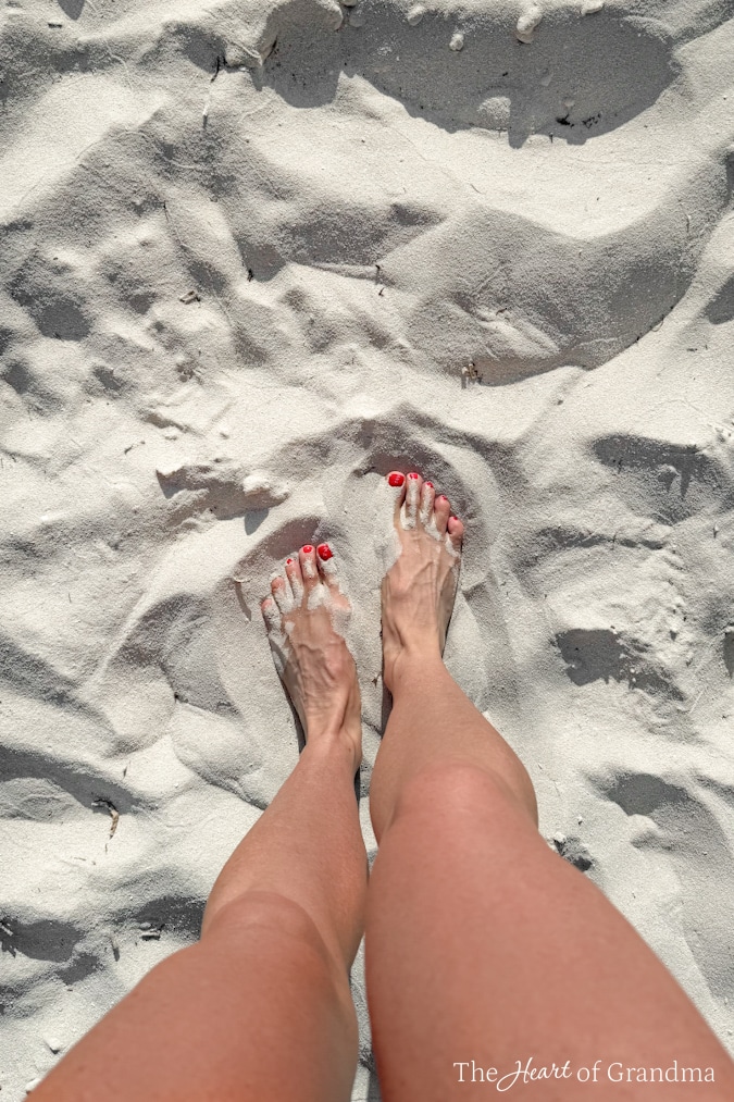 Two bare feet in white sand with red toenails peeking through.