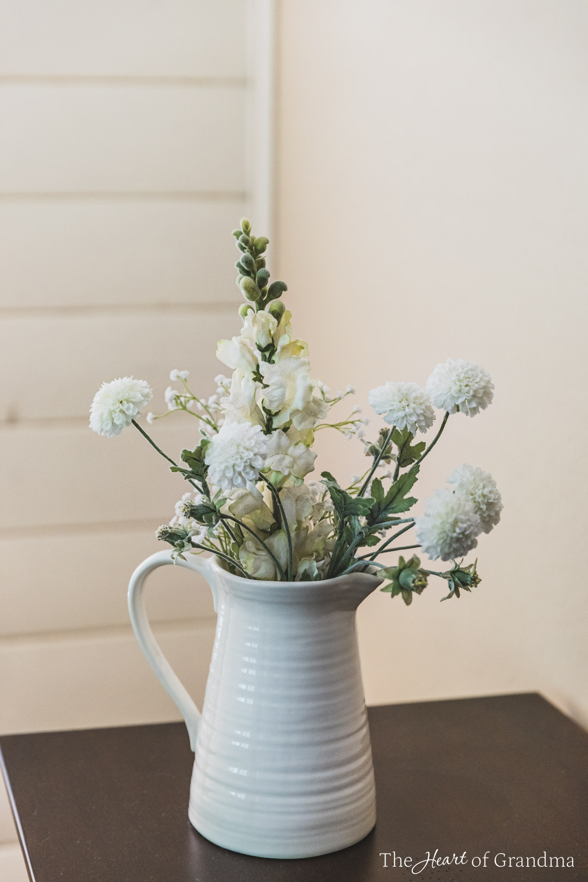 An arrangement of flowers in a white pitcher on the edge of a wooden dresser.