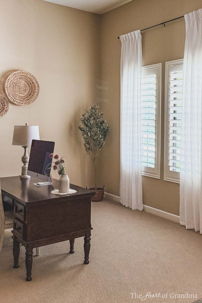 A wooden desk in front of a window with white drapes.