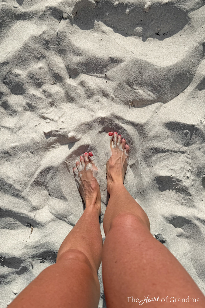 Two bare feet in white sand with red toenails peeking through.