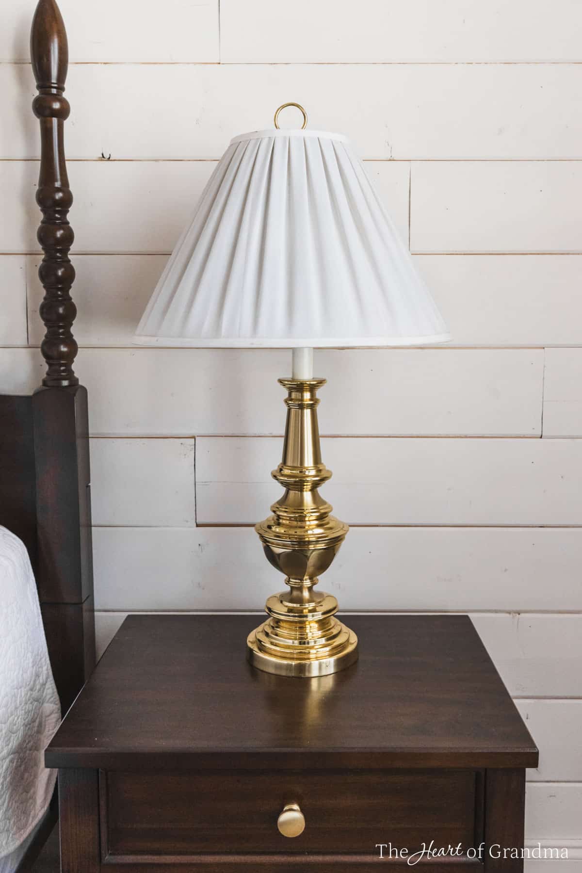 A solid brass lamp with a white pleated shade sitting on a nightstand.